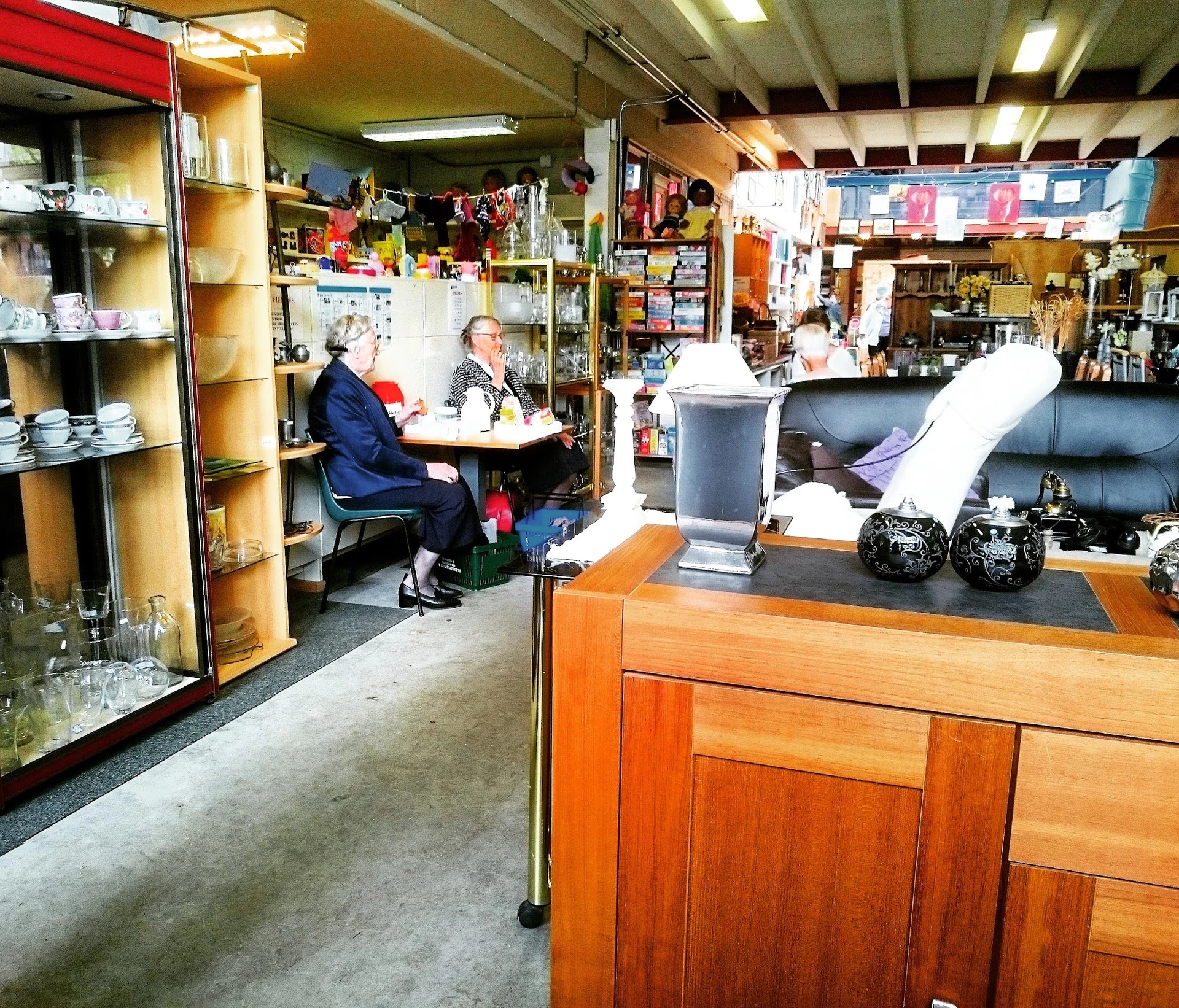 Winkelvloer van kringloopwinkel met houten toonbank, stellingen met spullen en bezoekers aan het winkelen