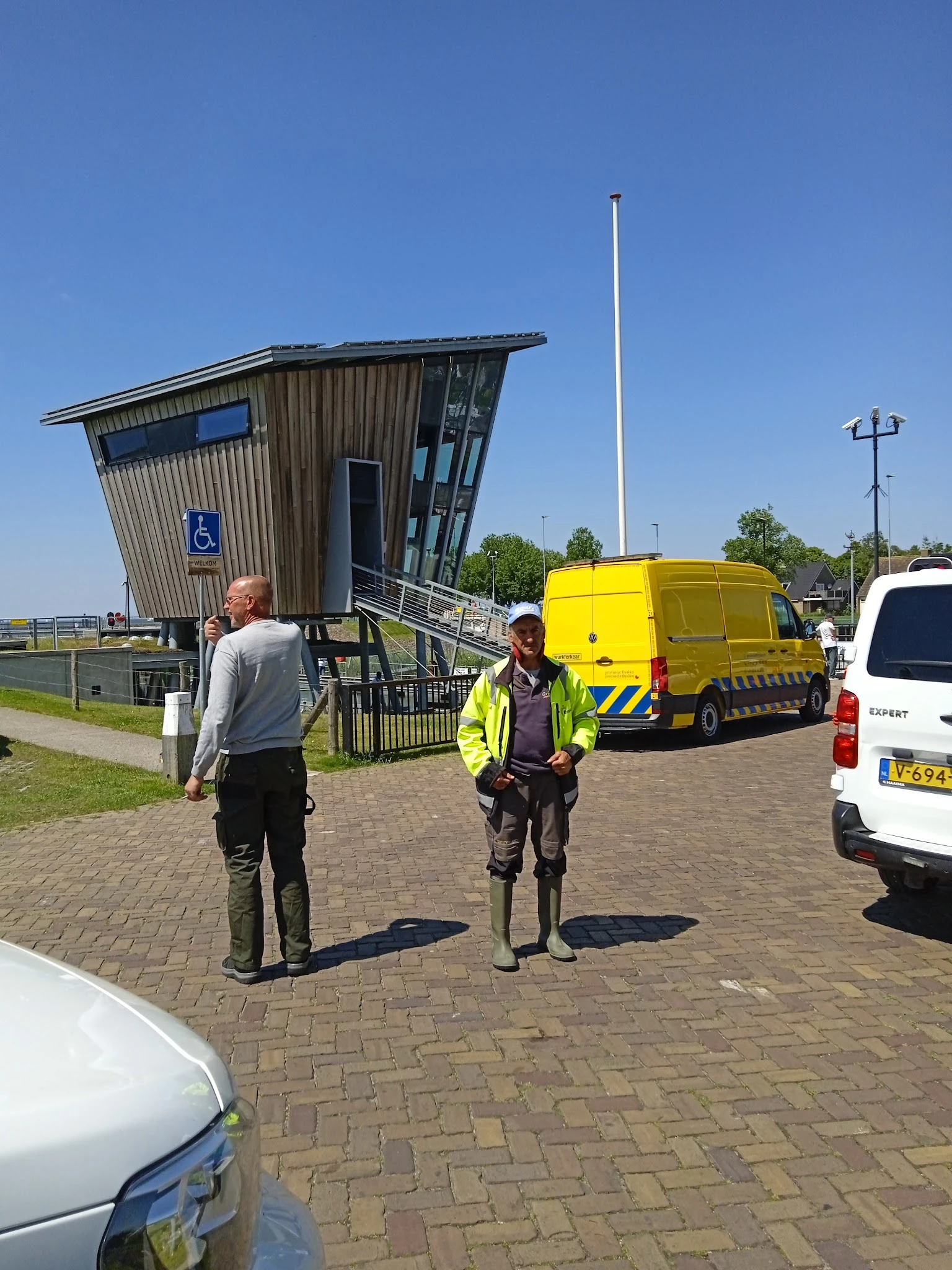 Twee personen in werkkleding en veiligheidsvesten staan buiten voor een scheef gebouwd gebouw met een gele bestelwagen