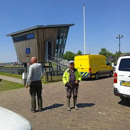 Twee personen in werkkleding en veiligheidsvesten staan buiten voor een scheef gebouwd gebouw met een gele bestelwagen