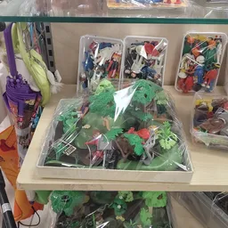 Uitgestalde speelgoedcollectie met actiefiguren, groene decoratieve planten en diorama's in plastic bakken op een tafel