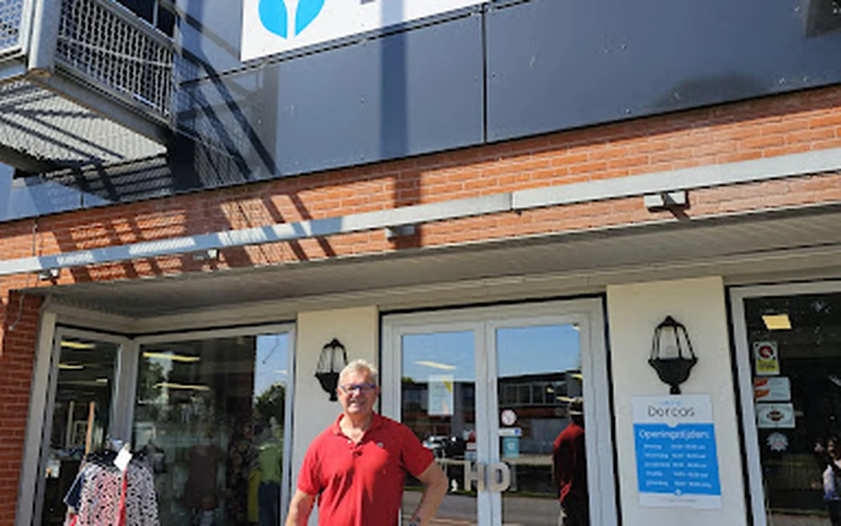 Voorgevel van kringloopwinkel Dorcas met glazen deuren, man in rood shirt ervoor, winkelnaam zichtbaar op gevel