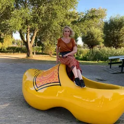 Vrouw zit op een geel speeltoestel in de vorm van een schoen op een plein met bomen