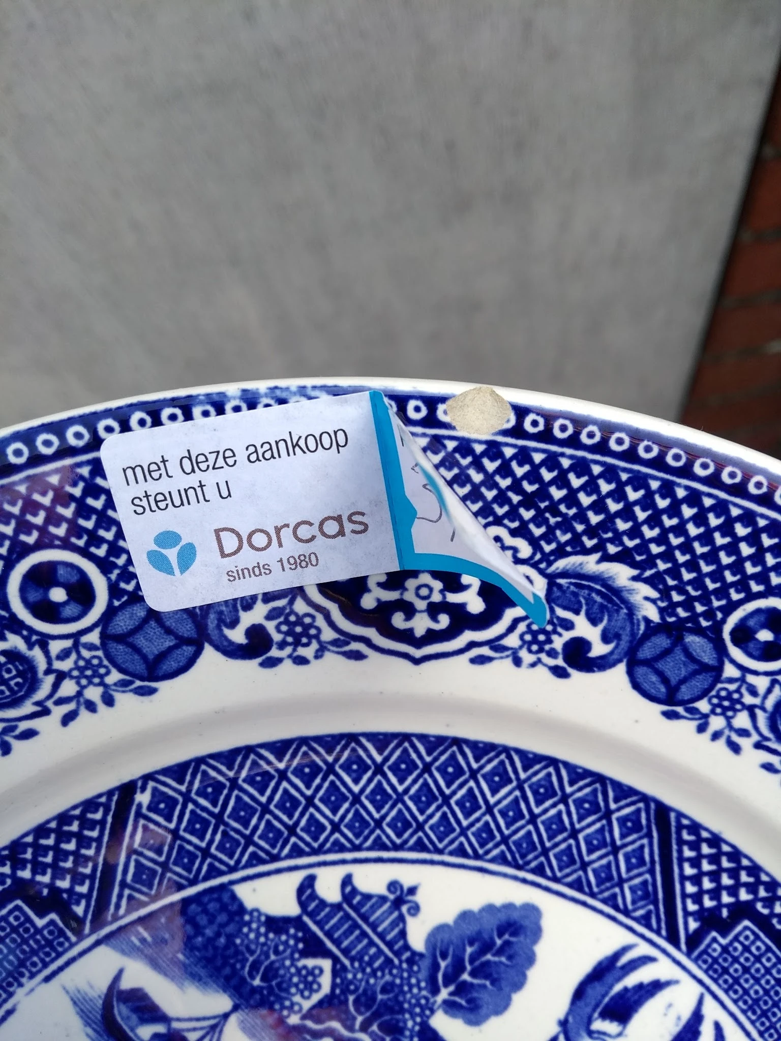 Blauw wit porselein schaaltje met Dorcas-label en Nederlandse tekst 'met deze aankoop steunt u Dorcas sinds 1980'