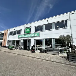 Witte gevel van kringloopwinkel Dromehof met grote ramen en groene letters op de voorgevel, gezien vanaf het parkeerterrein o