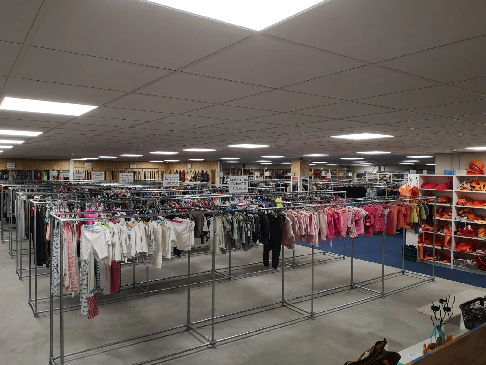Overzicht van de winkelruimte met rekken vol tweedehands kleding, goederen op planken en een heldere plafondverlichting