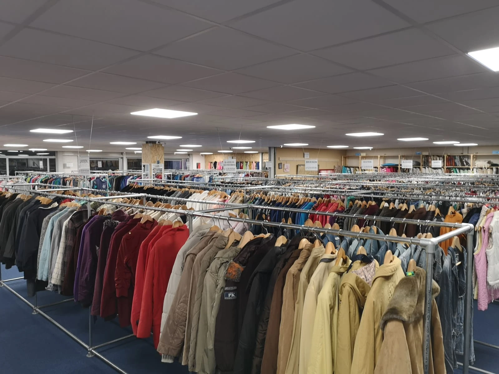 Lange rekken met tweedehands kleding in verschillende kleuren en stijlen, opgehangen aan houten hangers in een kringloopwinke