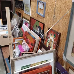 Kunstwerken en schilderijen gestapeld in en tegen een rek in de kringloopwinkel