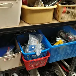 Stellingen met blauwe en rode plastic bakken en beige opbergmanden gevuld met diverse spullen in een kringloopwinkel