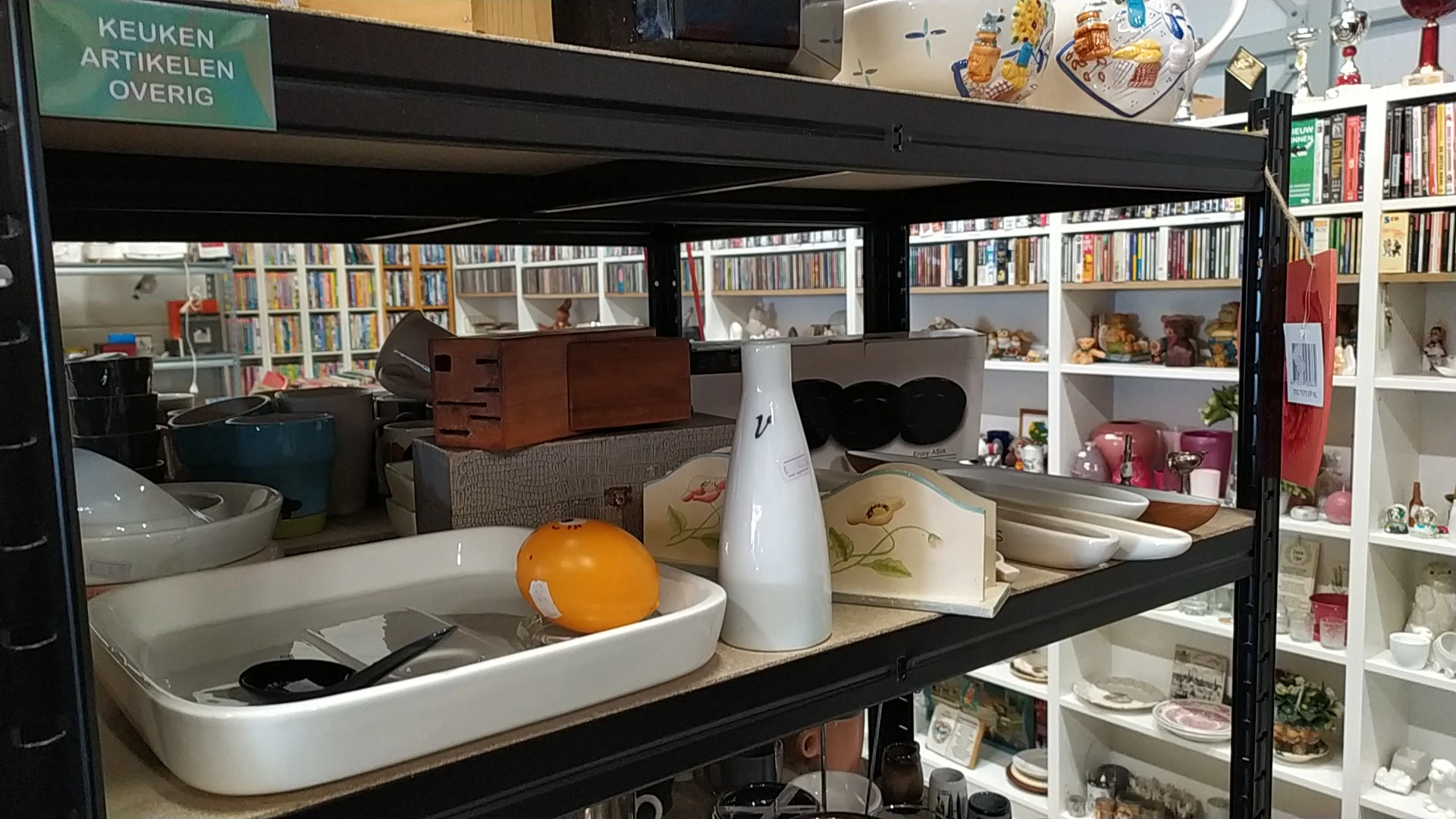 Glazen vitrine met huishoudelijke spullen: servies, een witte kan, decoratieve items en boeken op achtergrond