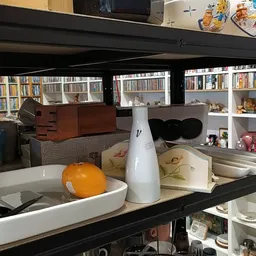 Glazen vitrine met huishoudelijke spullen: servies, een witte kan, decoratieve items en boeken op achtergrond