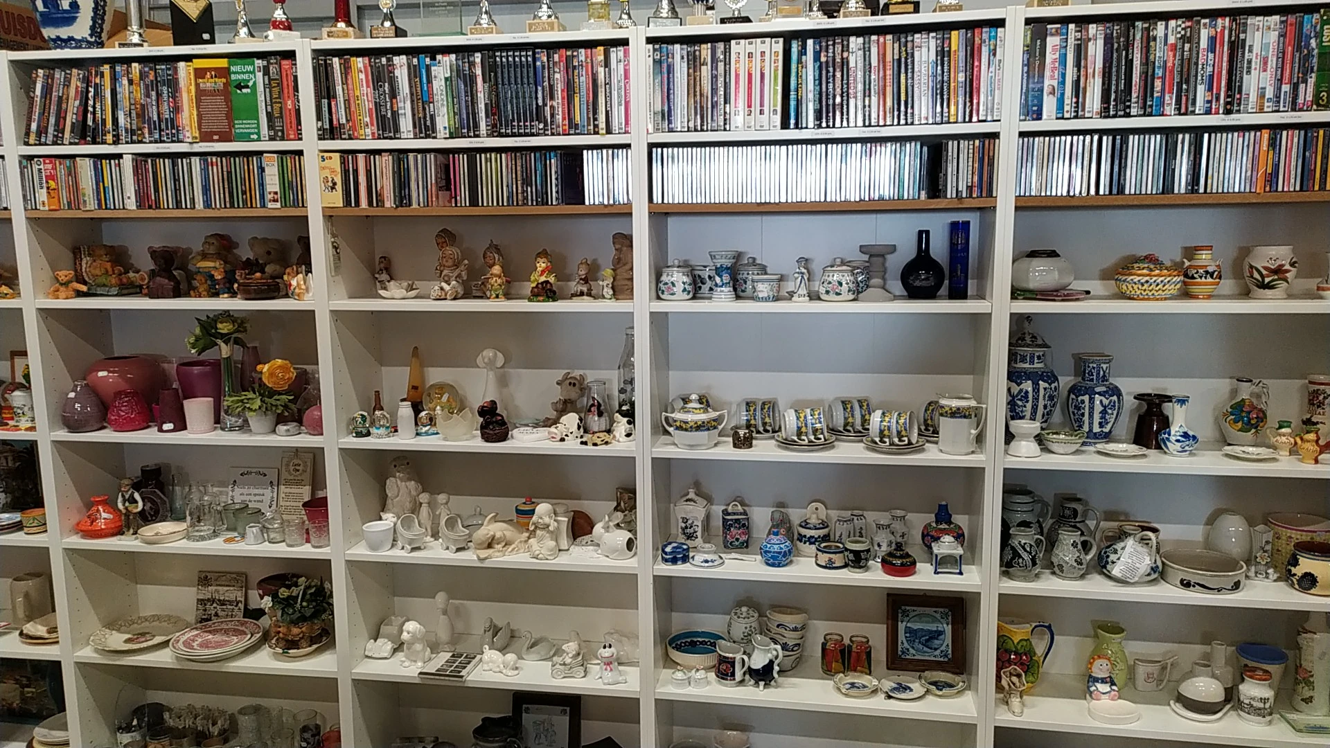 Witte boekenkast met dvd's, cd's bovenin en verzameling decoratieve items zoals beeldjes, servies en glazen op de planken ero