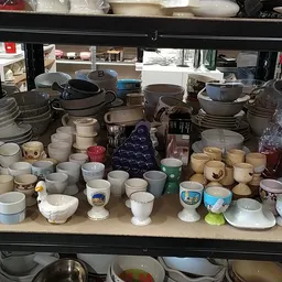 Uitgestalde serviesgoed en huishoudelijke artikelen op winkelrekken, waaronder kopjes, schalen, pannen en ander keramiek