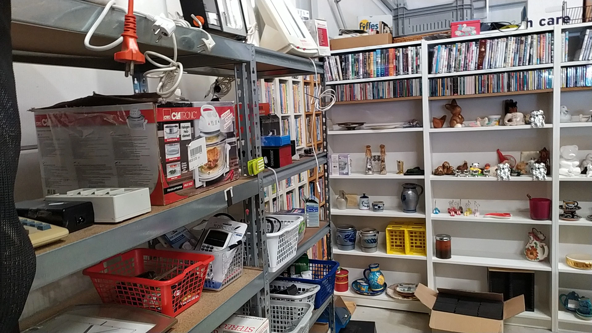Winkelbinnenzicht met stellingen vol dvd's, boeken en huishoudelijke spullen, rood en blauw winkelwagentjes