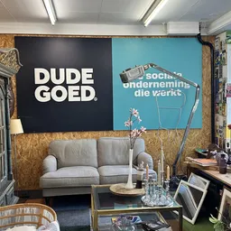 Winkelinterieur met grijze bank tegen muur met 'DUDE GOED' tekst, bureau, rekken met artikelen en winkelpui met daglicht