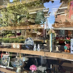 Vintage spullen in de etalage: servies, glazen, decoratieve objecten en een roze bloem op houten stellages achter glas