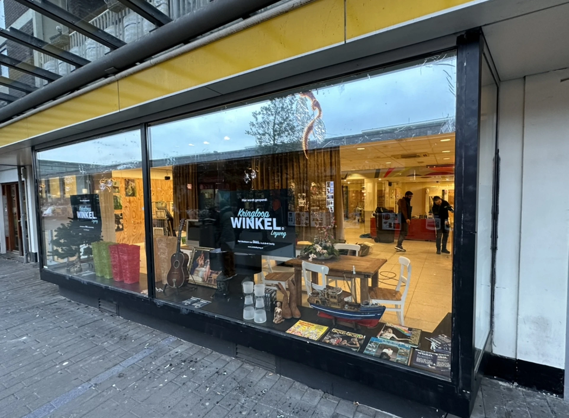 Winkelruit van kringloopwinkel met grote ramen, gele luifel, en zicht op het interieur met ingerichte spullen en meubels