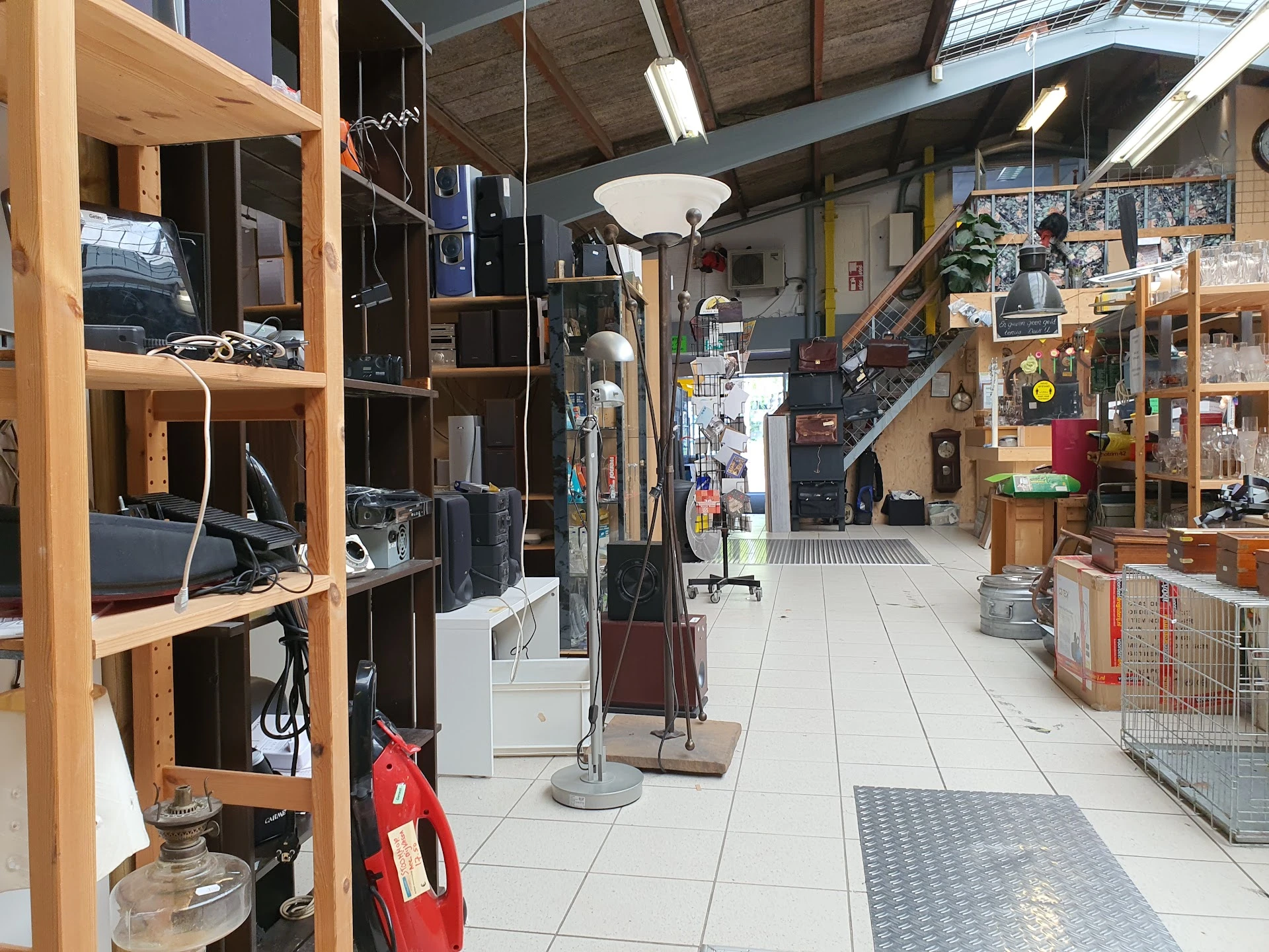 Binnenaanzicht van kringloopwinkel met houten stellingen, verlichtingsarmaturen, spullen op schappen en een brede doorgang