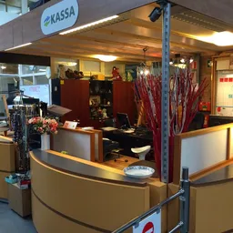 Kassahoek van kringloopwinkel met rood-geel interieur, personeel achter balie, uitzicht op winkelgedeelte met kleding