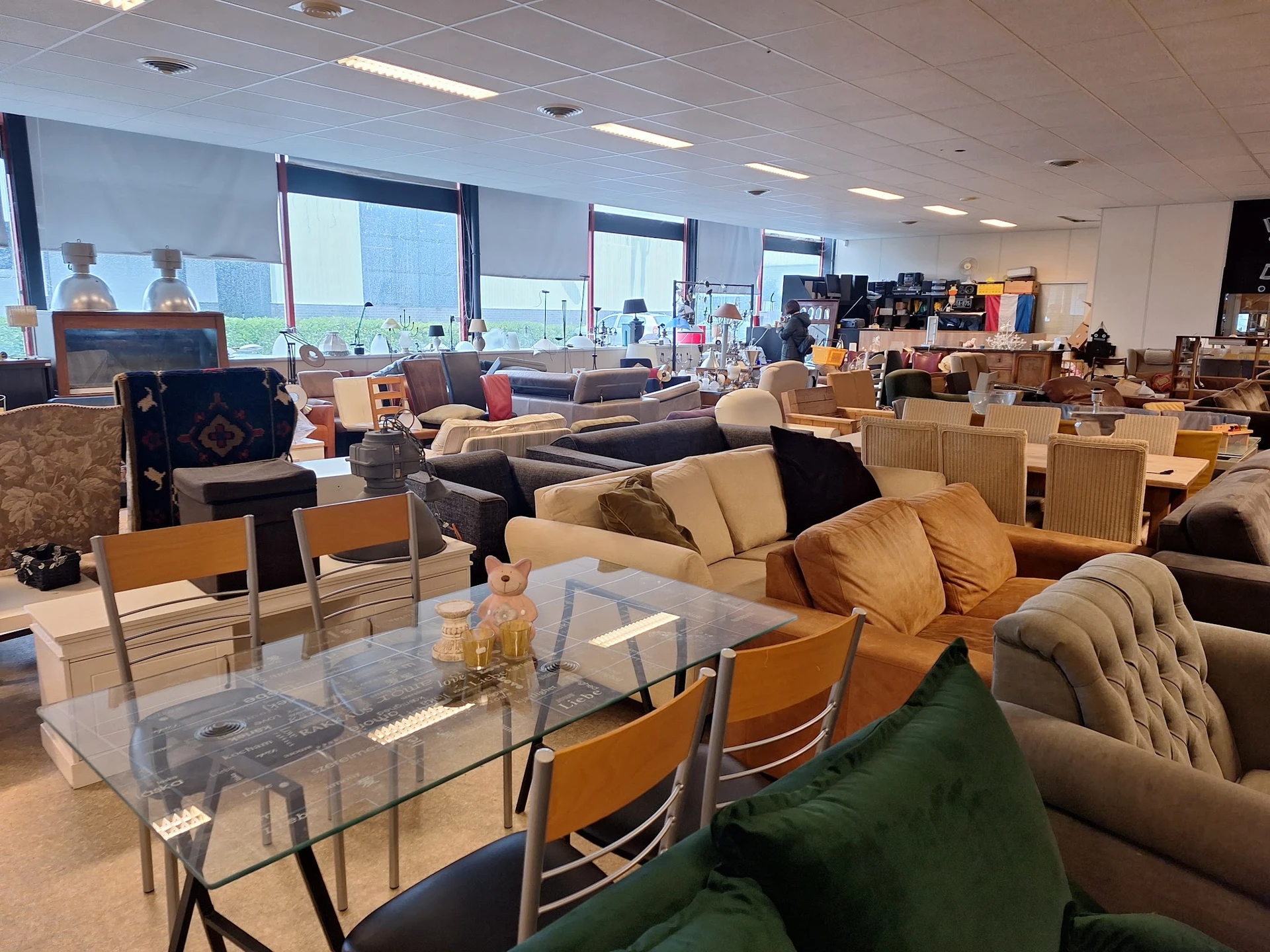 Inrichting van kringloopwinkel met meubels, zithoeken en fauteuils uitgestald in grote zaal met ramen.