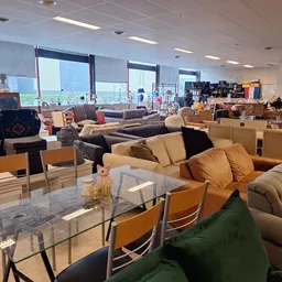 Inrichting van kringloopwinkel met meubels, zithoeken en fauteuils uitgestald in grote zaal met ramen.