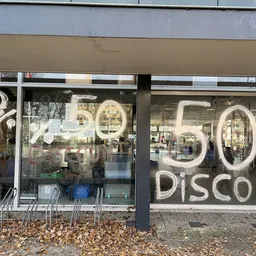 Winkelgevel met grote neonletters '50% DISCOUNT' op ramen, winkelnaam zichtbaar, enkele stoelen buiten