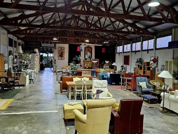 Ruim kringloopcentrum met diverse meubels, stoelen, tafels en huishoudartikelen uitgestald op betonnen vloer onder houten spa