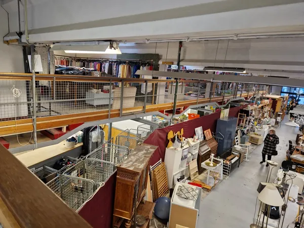 Overzicht van kringloopwinkel met meubelstukken, huishoudartikelen en dozen op de winkelvloer, galerijvloer boven zichtbaar