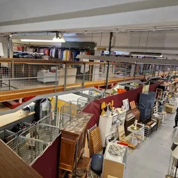 Overzicht van kringloopwinkel met meubelstukken, huishoudartikelen en dozen op de winkelvloer, galerijvloer boven zichtbaar