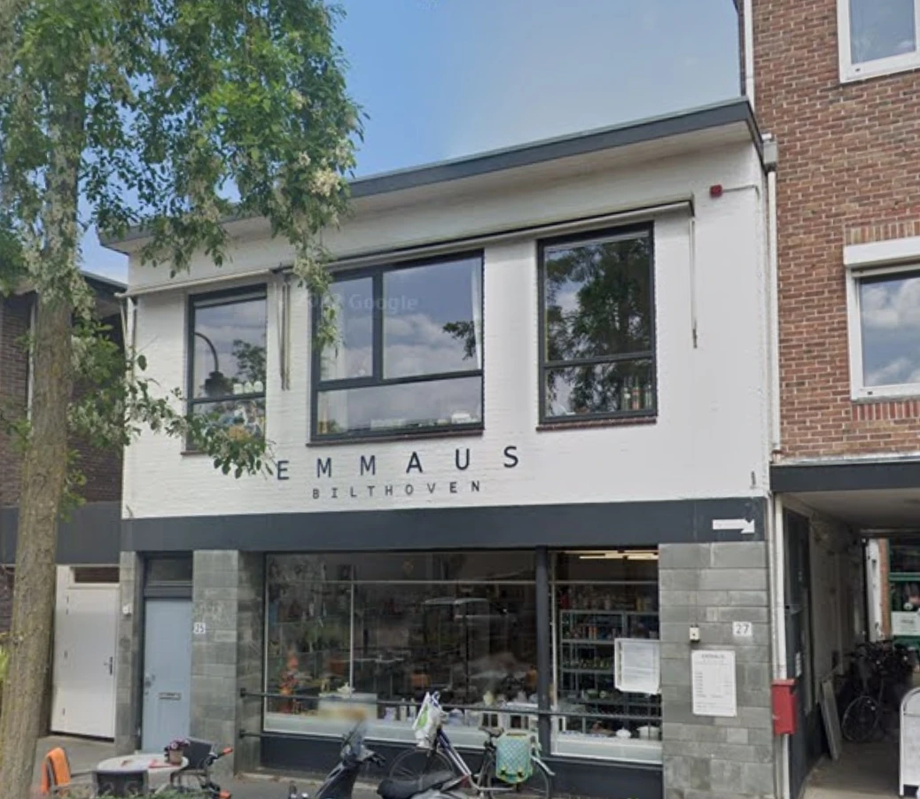 Witte gevel van kringloopwinkel Emmaus Bilthoven met grote ramen en donkere winkelpui.