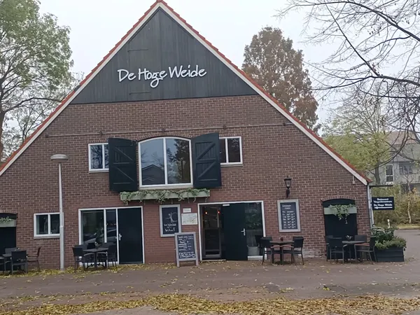 Voorgevel van bakstenen kringloopwinkel met gevel naam 'De Hoge Welde', ingang en terras met stoelen