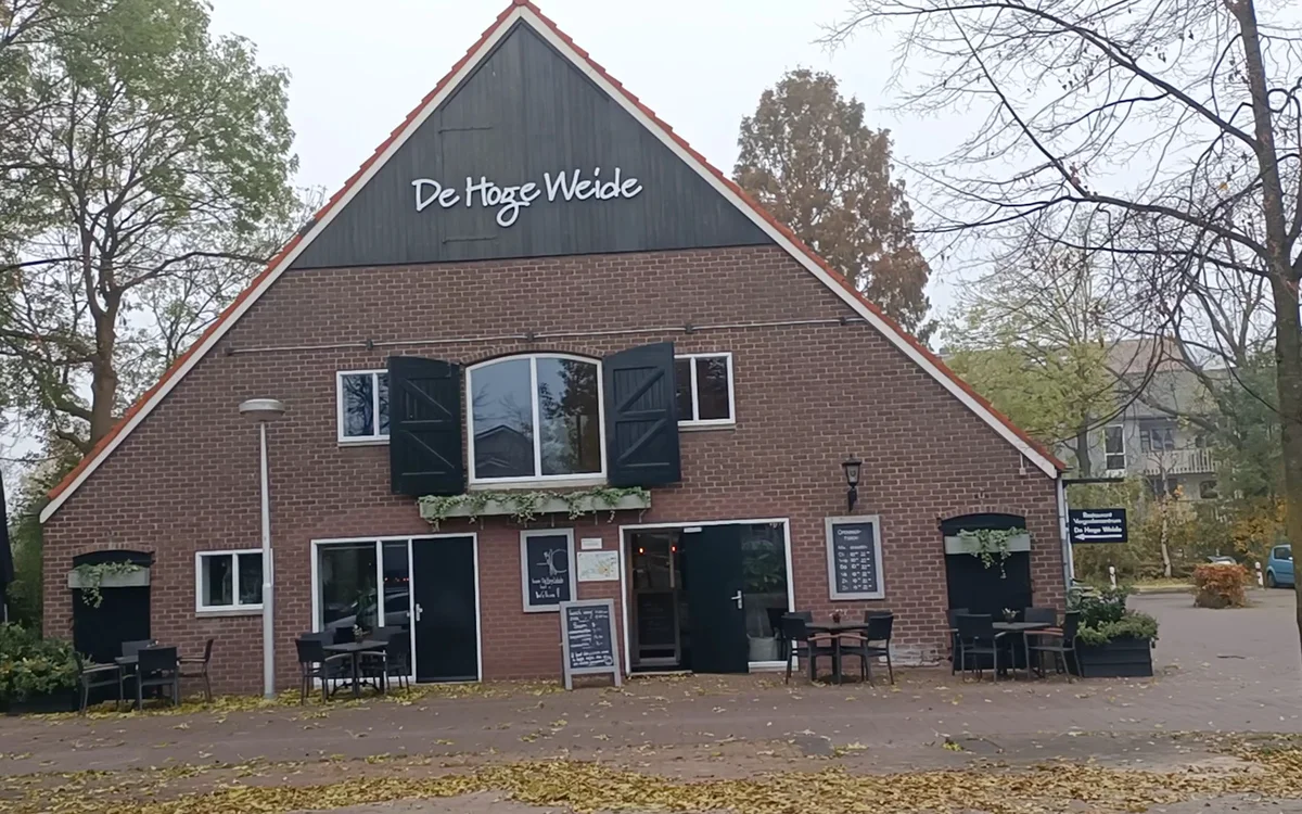 Voorgevel van bakstenen kringloopwinkel met gevel naam 'De Hoge Welde', ingang en terras met stoelen