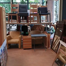 Tweedehands meubels en stoelen in de winkel, waaronder een oranje fauteuil, houten tafels en diverse stoelen.