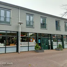 Gevel van kringloopwinkel met groene deuren, grote winkelramen en witte zijwanden, meerdere bovenramen