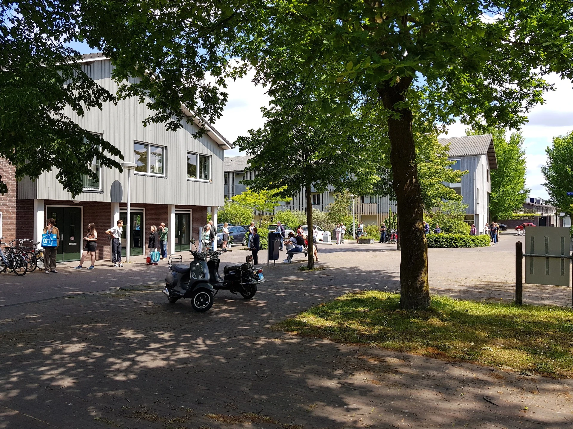 Voorkant van een moderne kringloopwinkel aan een plein met bomen, omgeven door voetgangers en fietsen