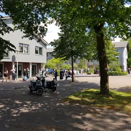 Voorkant van een moderne kringloopwinkel aan een plein met bomen, omgeven door voetgangers en fietsen
