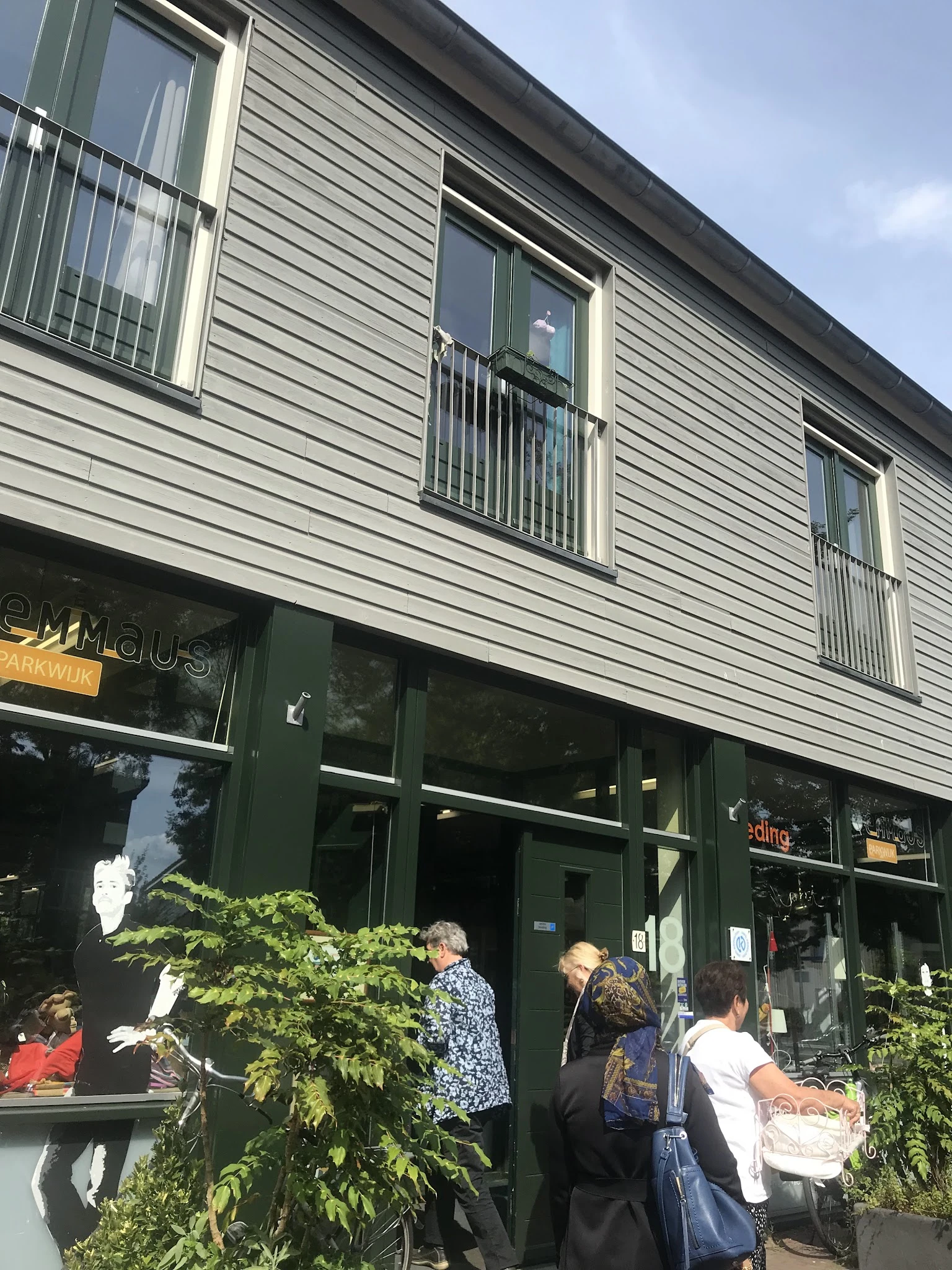 Gevel van kringloopwinkel met groene winkelruit en donkergroene deur, met enkele voorbijgangers voor de ingang