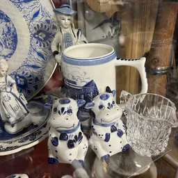 Blauwe en witte servies- en decoratieartikelen: kan, kom, decoratieve koeien en glas op een plank