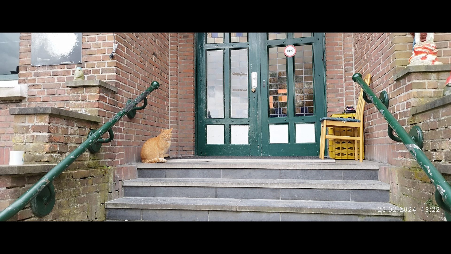 Ingang van een kringloopwinkel met groene deuren, rode bakstenen gevel en trappen, met een kat op de stoep
