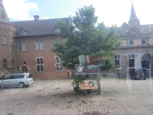 Bakstenen gevel van kringloopwinkel Emmaus Feniks met rood logo, omgeven door groenery, op een binnenplein