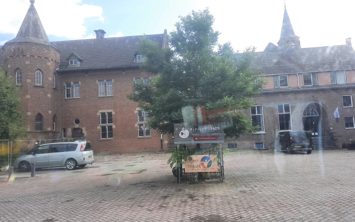 Bakstenen gevel van kringloopwinkel Emmaus Feniks met rood logo, omgeven door groenery, op een binnenplein