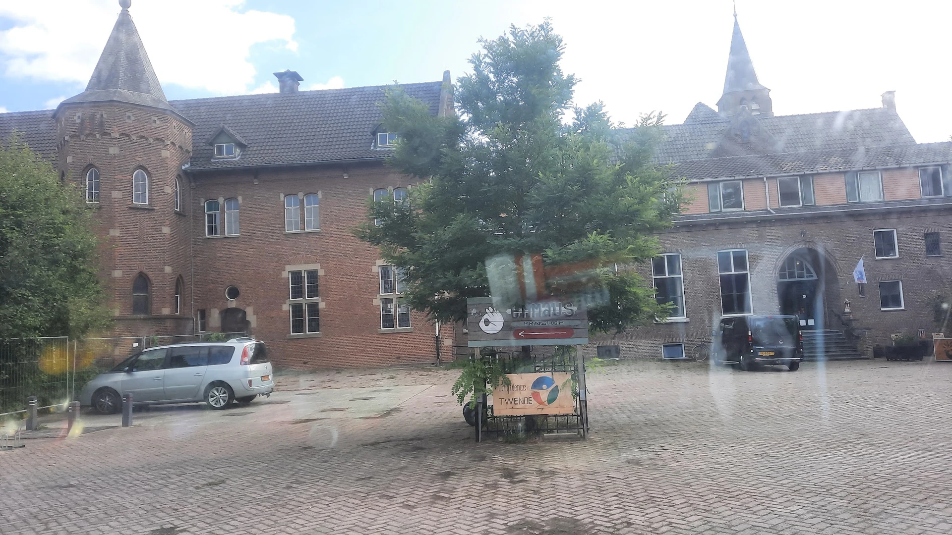 Bakstenen gevel van kringloopwinkel Emmaus Feniks met rood logo, omgeven door groenery, op een binnenplein