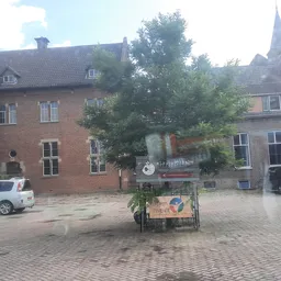 Bakstenen gevel van kringloopwinkel Emmaus Feniks met rood logo, omgeven door groenery, op een binnenplein