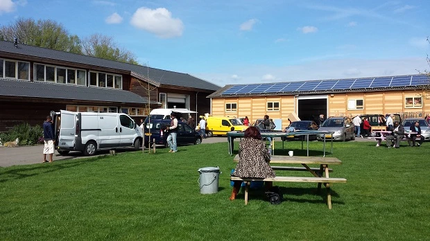 Groepsevenement op grasveld met picknickbanken, gebouwen en voertuigen op achtergrond