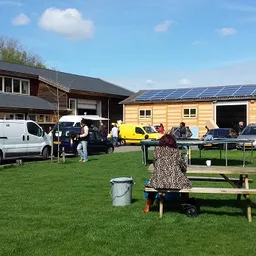 Groepsevenement op grasveld met picknickbanken, gebouwen en voertuigen op achtergrond