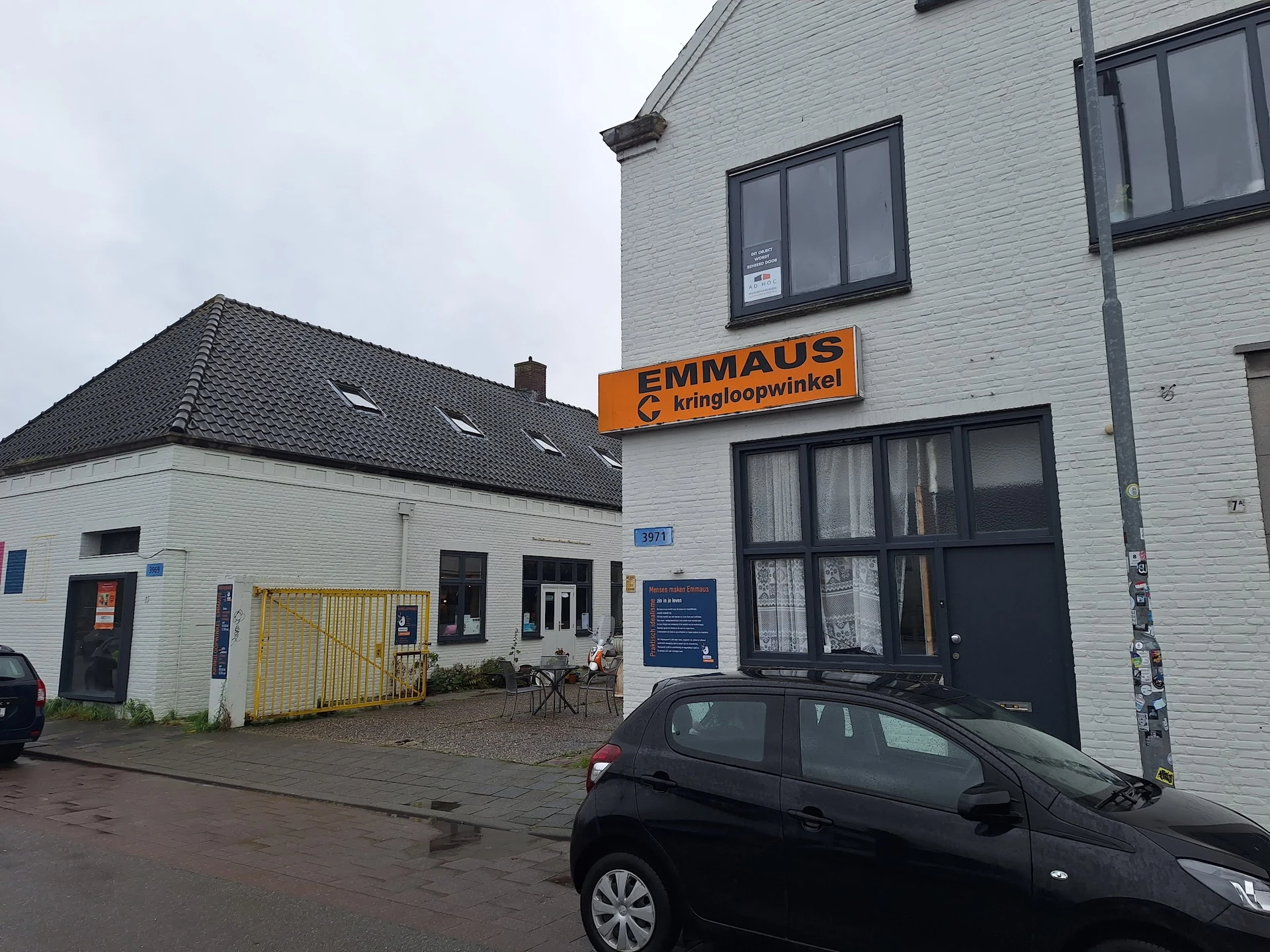 Voorgevel van Emmaus kringloopwinkel met oranje uithangbord, witte bakstenen gevel, ramen en parkeerterrein