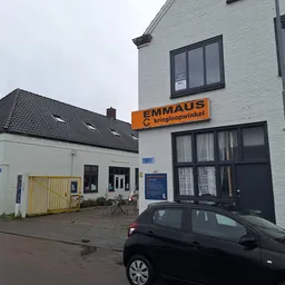 Voorgevel van Emmaus kringloopwinkel met oranje uithangbord, witte bakstenen gevel, ramen en parkeerterrein