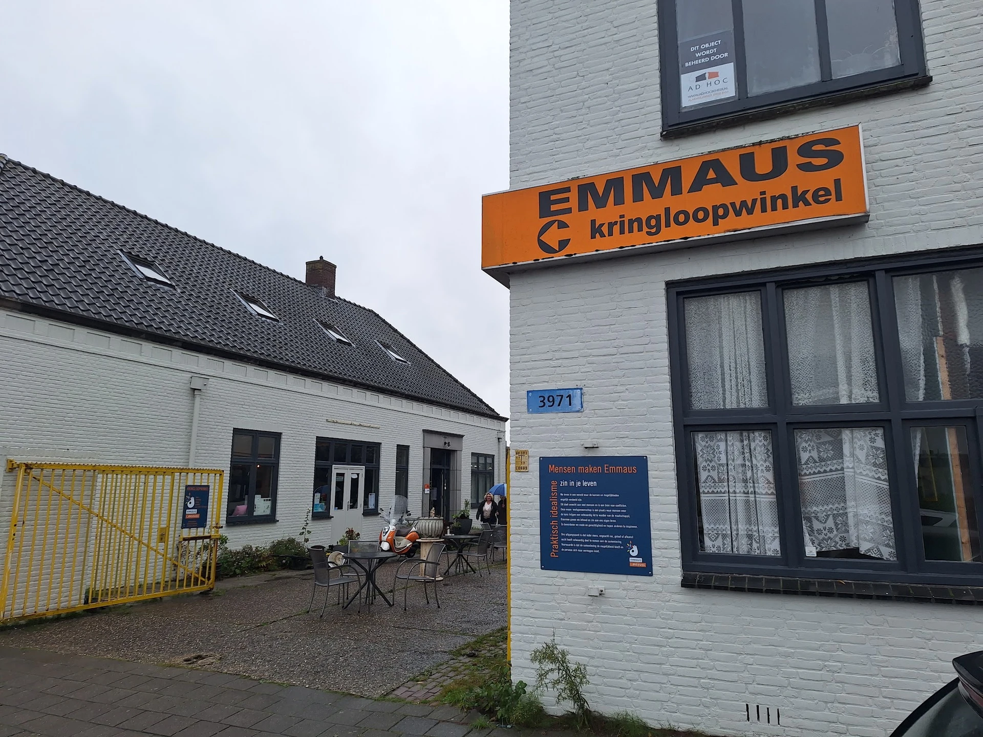 Voorgevel van kringloopwinkel met oranje logo, ingang en terras met stoelen