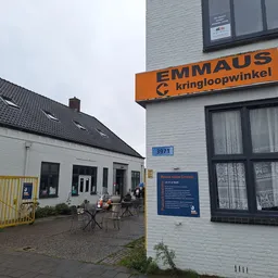 Voorgevel van kringloopwinkel met oranje logo, ingang en terras met stoelen