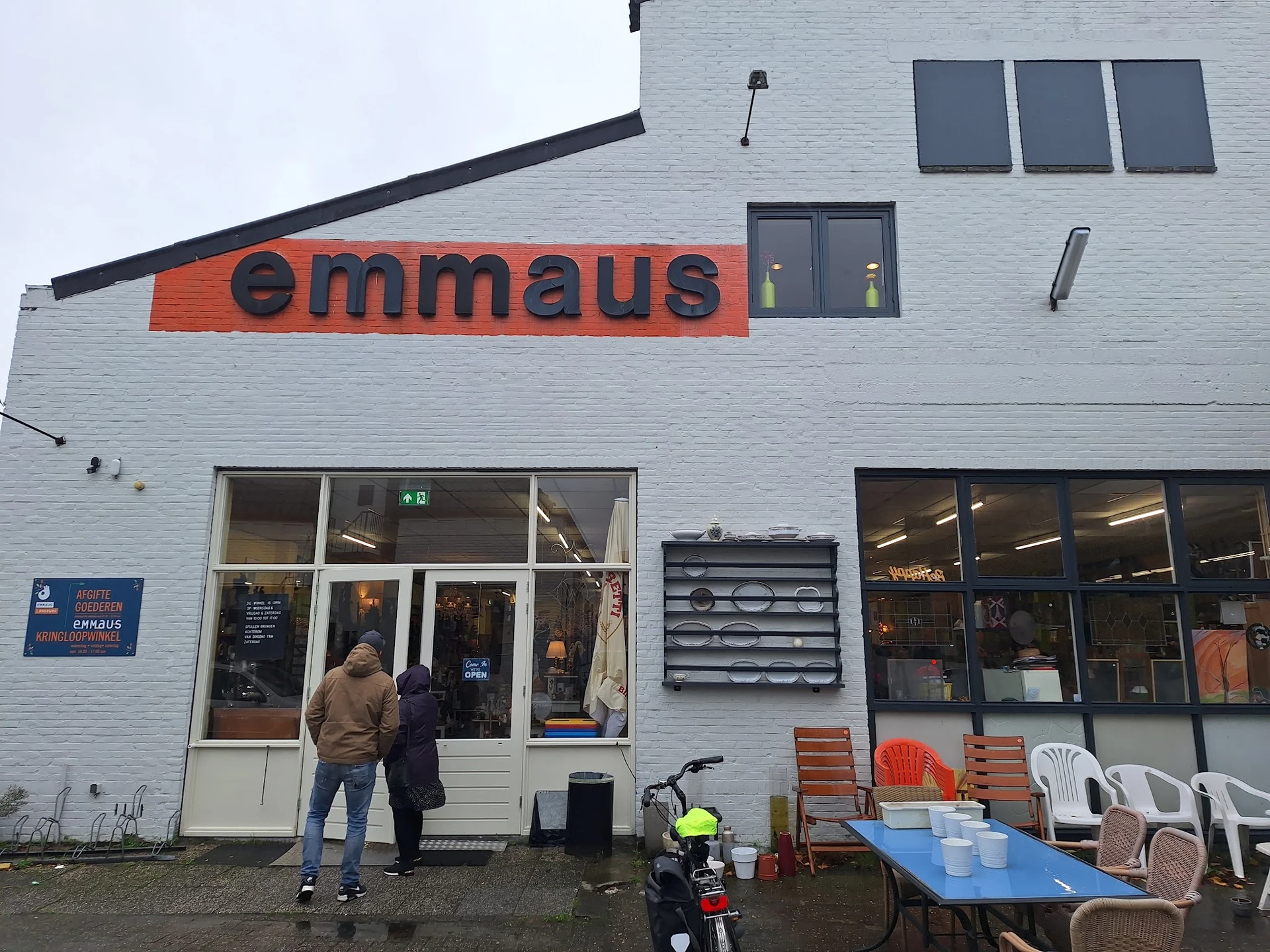 Witte gevel van kringloopwinkel met oranje 'emmaus' logo, ingang met glasdeuren en ramen, terras met stoelen ernaast