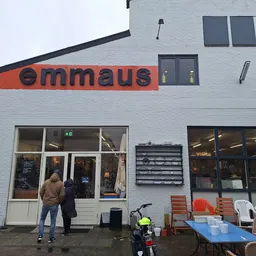 Witte gevel van kringloopwinkel met oranje 'emmaus' logo, ingang met glasdeuren en ramen, terras met stoelen ernaast